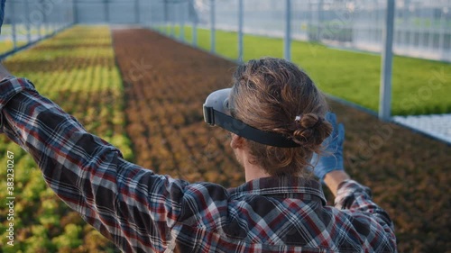 Farmer young engineer in VR glasses touching virtual user interface modificating plants and controlling water system in greenhouse. Organic farm business. New technologies. photo