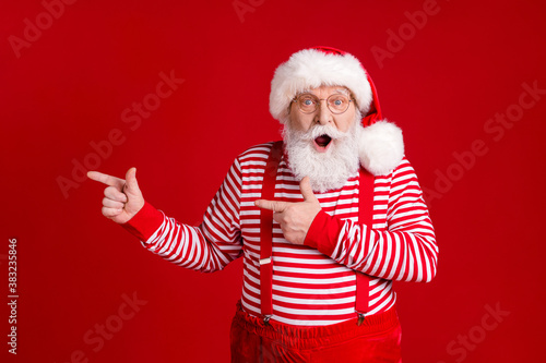Photo of retired old man white beard direct finger empty space shock present winter decoration wear santa x-mas costume suspenders spectacles striped shirt cap isolated red color background © deagreez