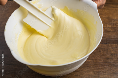 Folding smooth light yellow cake mixture with a spatula in a white bowl on top of wood table background.