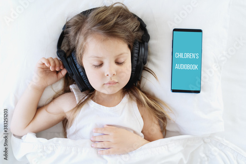 Fotografie Little girl listening to children's audiobook in mobile phone in headphones before sleeping in bed