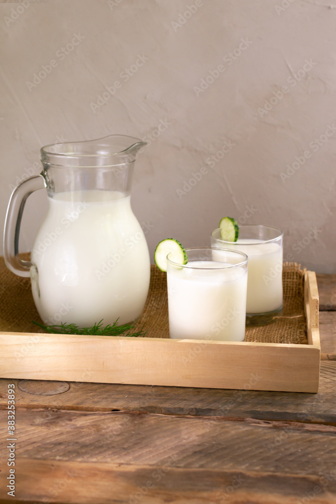 Two glasses with Ayran with a cucumber and a glass jug on a wooden tray on a wooden background. Fermented product concept. Copy space. Vertical orientation. Two glasses with Ayran with a cucumber and a glass jug on a wooden tray on a wooden background. Fermented product concept. Copy space. Vertical orientation.