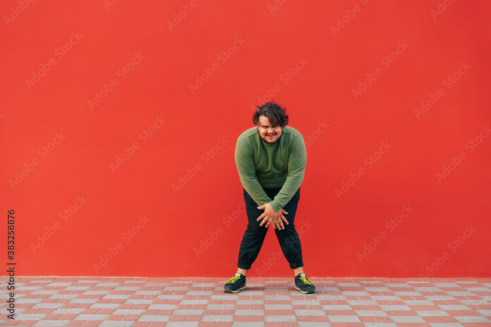 Funny fat man with curly hair dances on a red wall background and looks at the camera with a smile. Overweight man dancing on red background. Funny fat man with curly hair dances on a red wall background and looks at the camera with a smile. Overweight man dancing on red background.