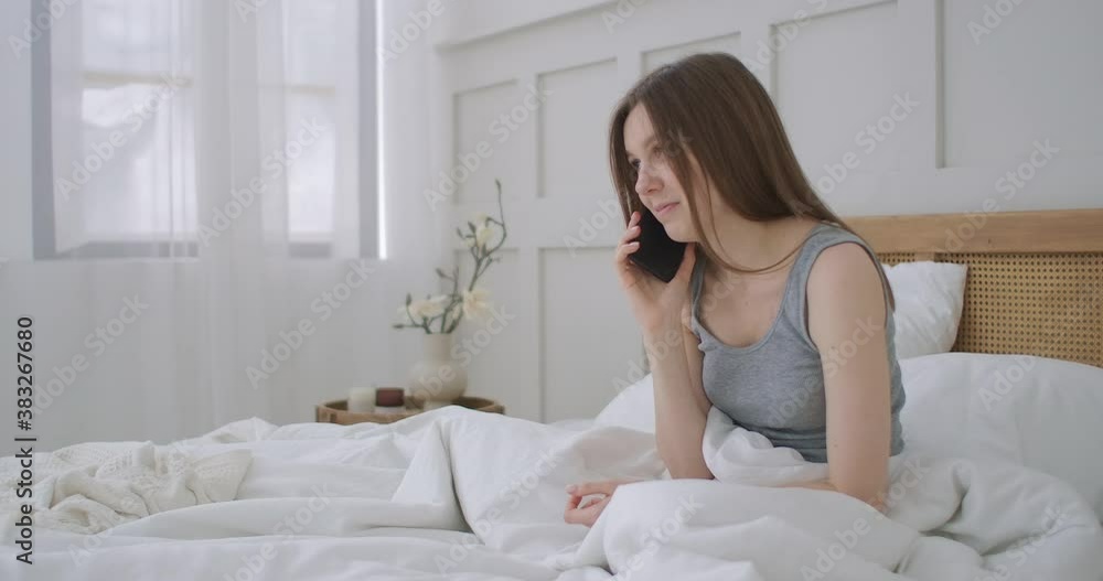 Young caucasian woman sitting on the bed in the morning feels bad. A young woman is making a smartphone call to her doctor. Young caucasian woman sitting on the bed in the morning feels bad. A young woman is making a smartphone call to her doctor.
