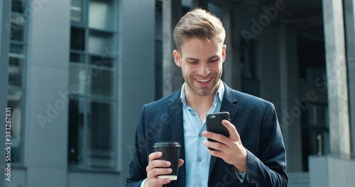 Handsome Successful Businessman is smiling Charmingly while browsing Internet. Happy Caucasian Man is using Smartphone outdoor in Business District, holding Coffee to go. Wearing stylish Suit. photo