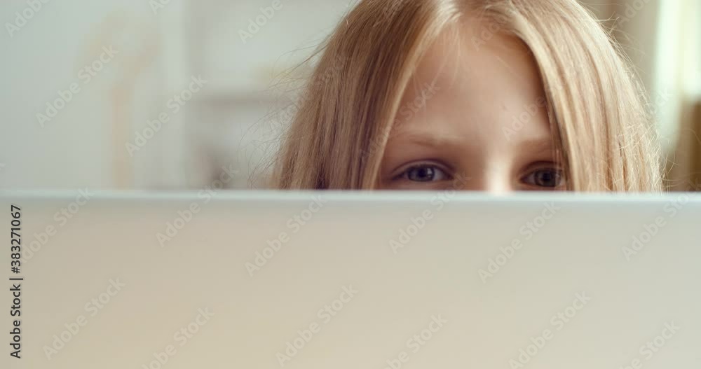 Portrait of little cute girl hiding behind laptop while sitting at table. Close-up of female blue eyes of child peeking out from behind computer screen, plays during class, indulges in doing homework Portrait of little cute girl hiding behind laptop while sitting at table. Close-up of female blue eyes of child peeking out from behind computer screen, plays during class, indulges in doing homework