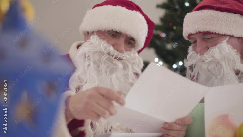 Young and old Santas examining letters on Christmas eve. Portrait of two men in red Christmas costumes reading wishes on New Year holidays. Young and old Santas examining letters on Christmas eve. Portrait of two men in red Christmas costumes reading wishes on New Year holidays.