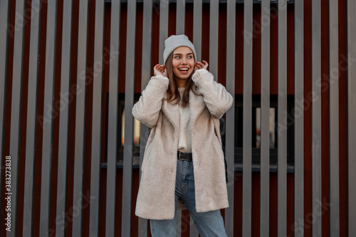 Trendy cheerful young woman straightens stylish gray knitted hat outdoors.Attractive happy girl with a cute smile in a fashionable faux fur coat in jeans posing near a metal vintage wall in the street photo