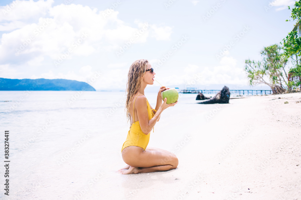Side view of Caucasian woman in yellow swimsuit drinking coconut beverage during sunbathing on tropical Hawaii beach, young female enjoying healthy vegan food during summer vacations on Seychelles