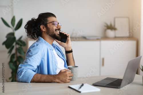 Cheerful Indian Freelancer Guy Resting With Coffee And Cellphone At Home Office