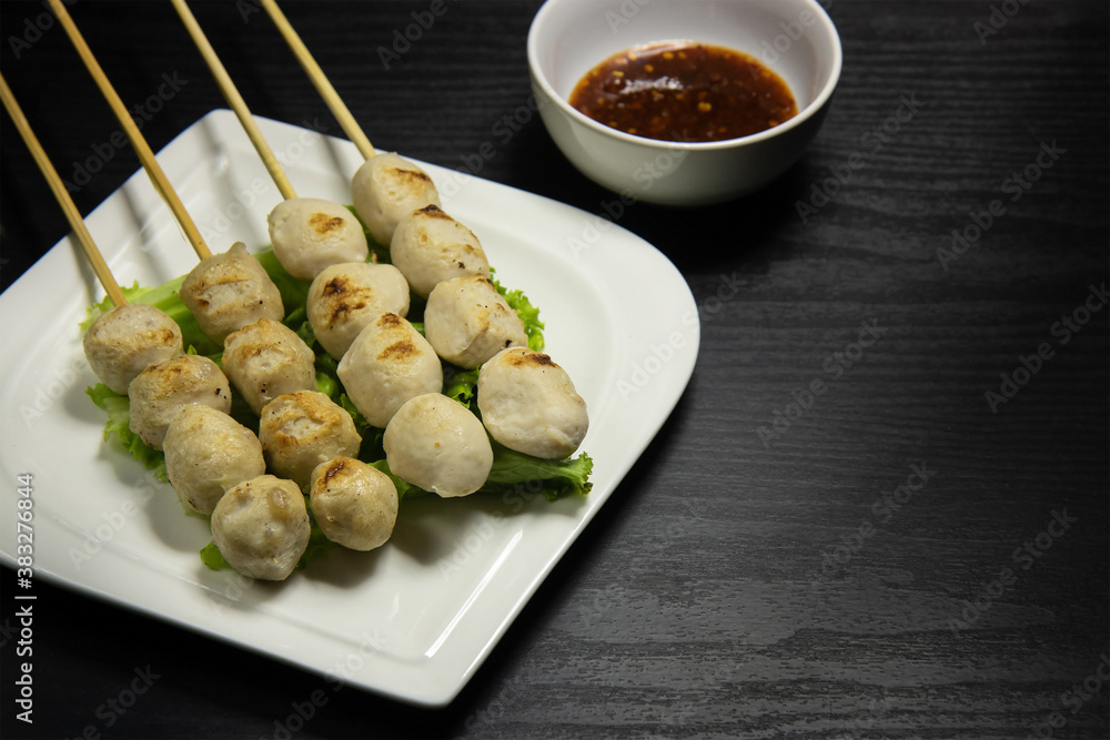 Grilled pork meatballs skewer in a white plate and sauce in a white bowl on a black background. Grilled pork meatballs skewer in a white plate and sauce in a white bowl on a black background.