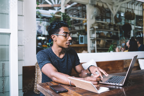 Serious Hispanic businessman taking notes while using laptop in cafe