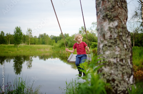 Little boy having fun on a swing hanging on big tree near pond or river in the forest in summer day.