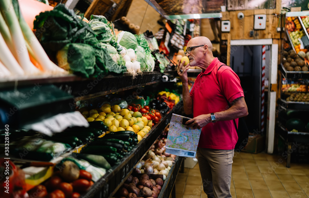 Obraz premium Elderly Caucasian pensioner with location map in hand enjoying smell of fresh lemon during shopping time for buying veggie vitamins, aged male tourist choosing fruits and vegetables in fruteria