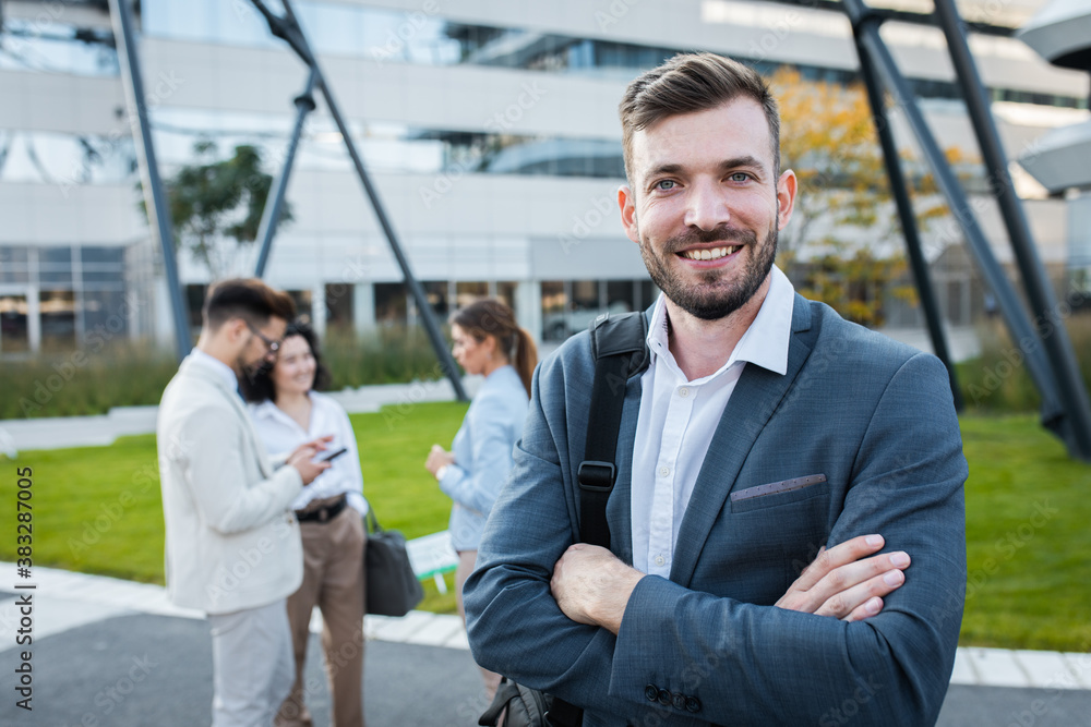 Portrait of smiling businesswoman standing in front of modern office buildings with colleagues behind him.