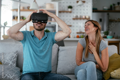 Smiling young man using VR headset at home on couch. Woman and her husband enjoying virtual reality in apartment..