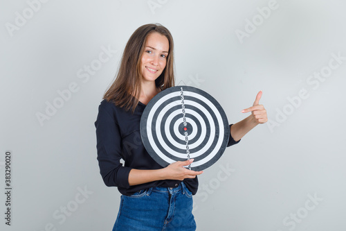 Young woman in black shirt, jeans shorts pointing finger at dartboard and looking hopeful , front view.