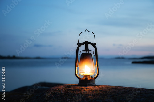 Large oil lamp outdoors in the dark. Old lantern illuminates on the sea shore. Rocks and sunset sky. © raland