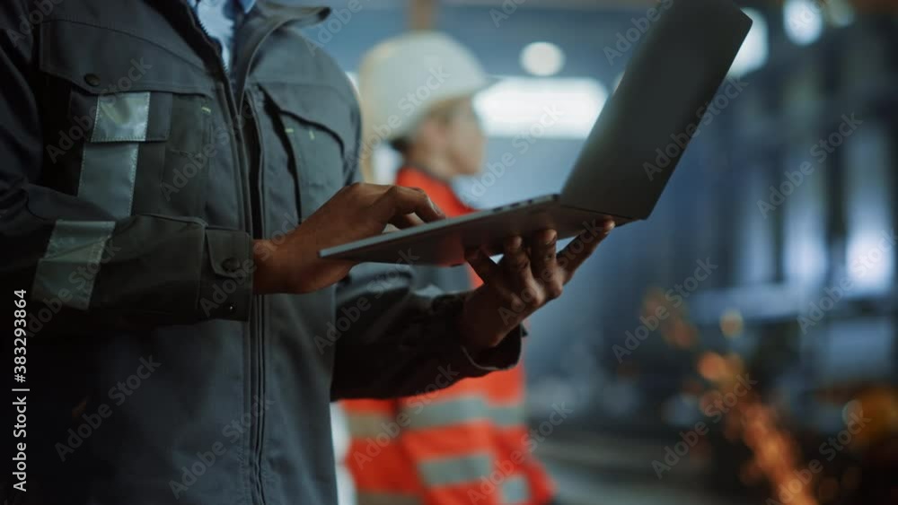 Close Up on Hands of a Professional Heavy Industry Engineer Wearing Safety Uniform and Using Laptop Computer. African American Industrial Specialist Standing in a Metal Construction Manufacture.