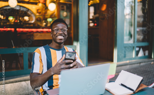 Cheerful black man with smartphone and laptop in street cafe on summer terrace