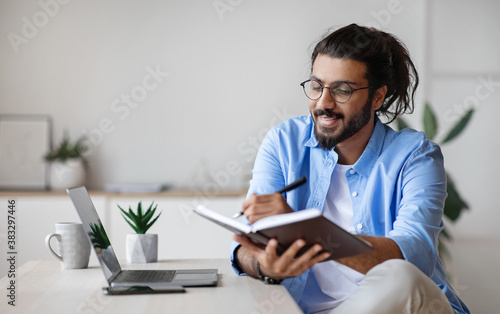 Smiling Western Freelancer Guy Taking Notes At Workplace In Home Office