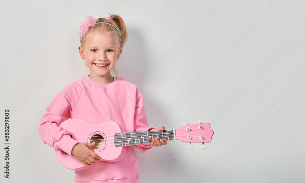Cute caucasian little girl 4-5 year old in pink sweatshirt are playing the ukulele against gray studio background