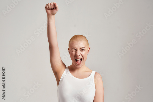 Portrait of a beautiful young caucasian woman with shaved head wearing white shirt, winking at camera and raising clenched fist, standing isolated over grey background photo