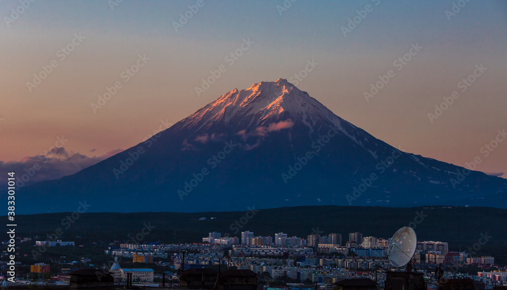 View of the volcano at sunset. The top of the volcano is illuminated by the sun. In the foreground is the evening city