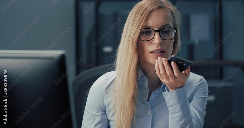 Successful woman in eyewear having business talk on smartphone while working on modern computer at office. Concept of communication and technology. Successful woman in eyewear having business talk on smartphone while working on modern computer at office. Concept of communication and technology.