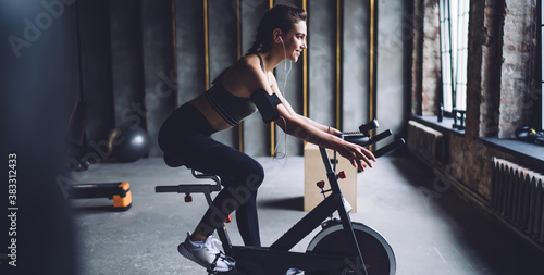 Smiling caucasian female in activewear sitting on exercising bike on cardio workout listening music, cheerful woman satisfied with sound accessory entertaining during training with playlist songs