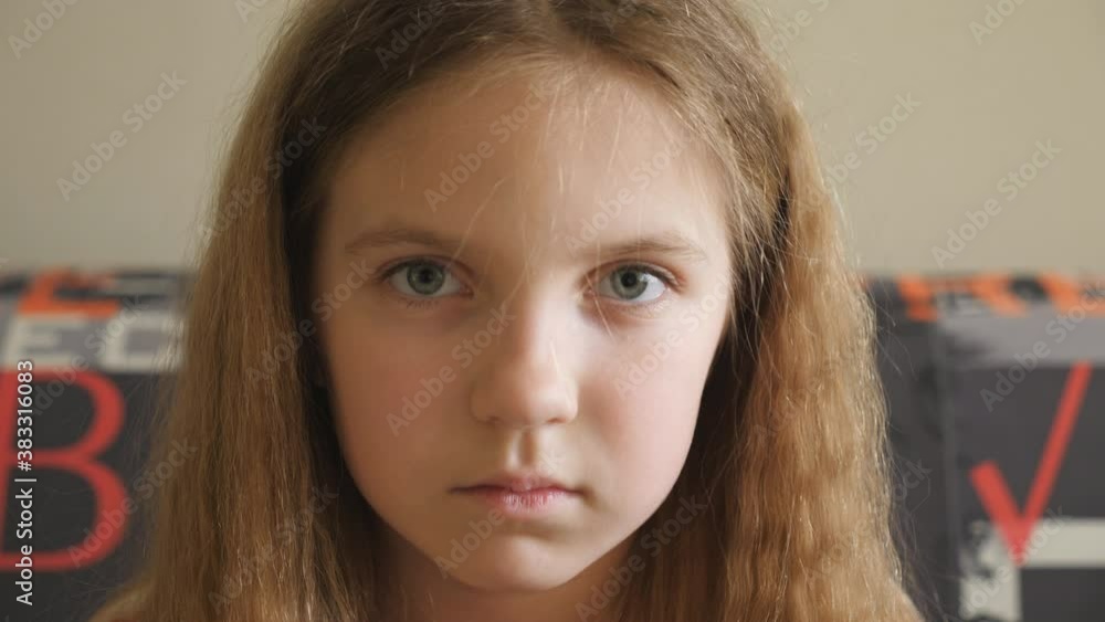 Small disappointed kid sitting on the couch and looking into camera. Portrait of unhappy little girl with blonde hair indoor. Close up emotions of female child with sorrowful expression on her face Small disappointed kid sitting on the couch and looking into camera. Portrait of unhappy little girl with blonde hair indoor. Close up emotions of female child with sorrowful expression on her face