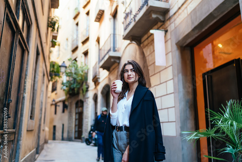 Pensive female tourist in trendy coat standing at street in historic center and pondering on old times, Caucasian hipster girl with disposable takeaway cup thinking during travel sightseeing