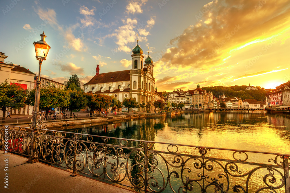 Colorful sunset in Lucerne city on Lake Lucerne in Switzerland. Jesuitenkirche or Church of St. Francis Xavier reflects on Reuss river. Pedestrian bridge in liberty style balcony and iron street lamps