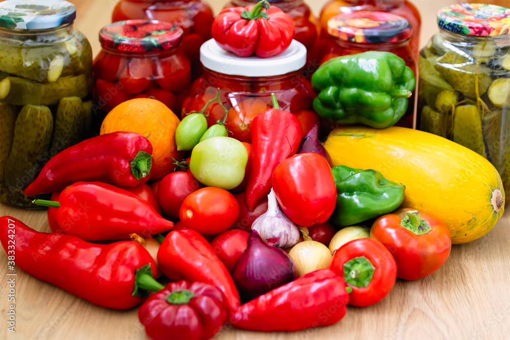 Canned cucumbers and tomatoes in jars on a wooden background. Lots of fresh peppers and tomatoes. Preservation of vegetables for the winter at home. Canned cucumbers and tomatoes in jars on a wooden background. Lots of fresh peppers and tomatoes. Preservation of vegetables for the winter at home.