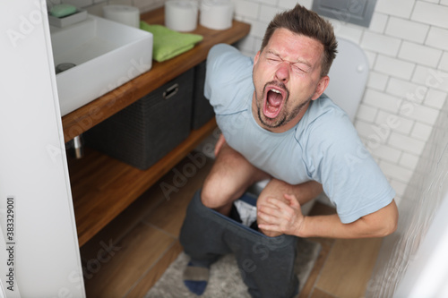 Young man with suffering on his face sits on toilet in bathroom. Indigestion constipation concept. photo