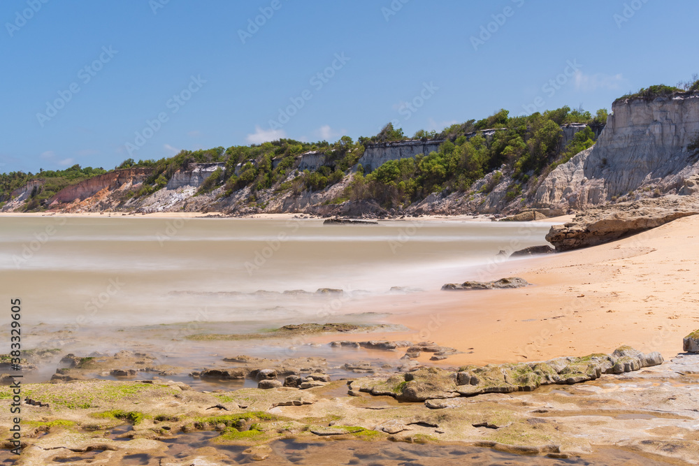 Tropical beach with reefs, yellow sand, silky sea and clear cliffs on a beautiful blue sky day.