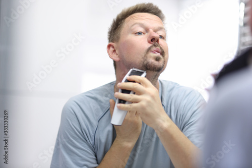Young man shaves his face with razor while looking in mirror in bathroom. Daily morning hygiene procedures for men concept.
