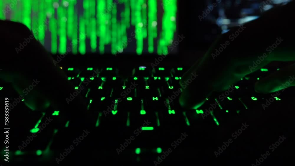 Cybersecurity concept. Hacker in dark room entering code on green lit keyboard, hacker code in the background Cybersecurity concept. Hacker in dark room entering code on green lit keyboard, hacker code in the background