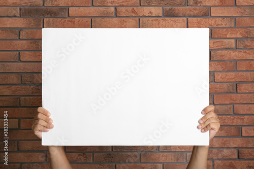 Man holding white blank poster near red brick wall, closeup. Mockup for design