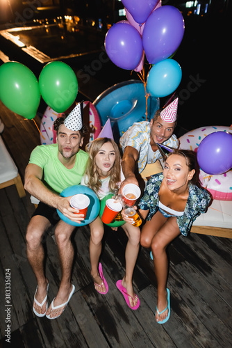 Young friends looking at camera while toasting with beer near balloons and swimming pool at night