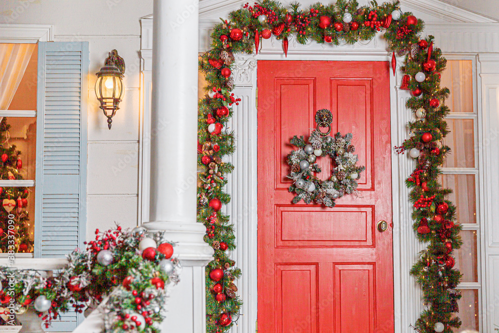 Christmas porch decoration idea. House entrance with red door decorated for holidays. Red and green wreath garland of fir tree branches and lights on railing. Christmas eve at home Christmas porch decoration idea. House entrance with red door decorated for holidays. Red and green wreath garland of fir tree branches and lights on railing. Christmas eve at home