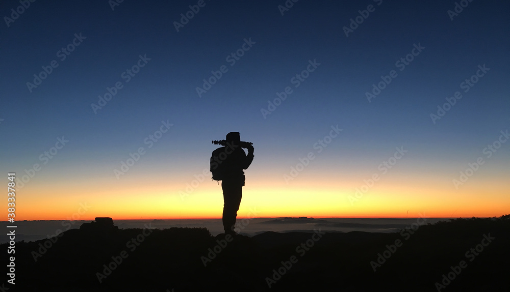 Silhouette of an adventurous backpacker traveler holding a tripod on top of the mountain at sunset or sunrise.