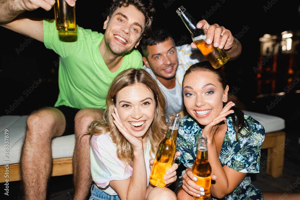 Selective focus of women with beer bottles looking at camera near boyfriends at night Selective focus of women with beer bottles looking at camera near boyfriends at night