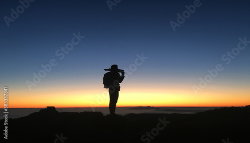 Silhouette of an adventurous backpacker traveler holding a tripod on top of the mountain at sunset or sunrise.