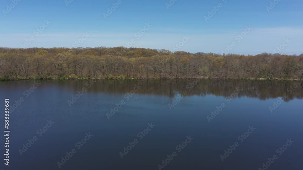 An aerial view of a blue, reflective lake with trees lining the lake's edge. The sky is blue & clear. The drone slowly boom up as it dolly in and pan right to the trees reflecting onto the waters. An aerial view of a blue, reflective lake with trees lining the lake's edge. The sky is blue & clear. The drone slowly boom up as it dolly in and pan right to the trees reflecting onto the waters.