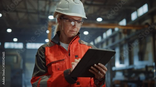 Professional Heavy Industry Engineer/Worker Wearing Safety Uniform and Hard Hat Uses Tablet Computer. Serious Successful Female Industrial Specialist Walking in a Metal Manufacture Warehouse. photo