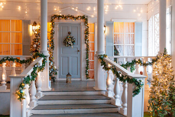 Christmas porch decoration idea. House entrance decorated for holidays. Golden and green wreath garland of fir tree branches and lights on railing. Christmas eve at home