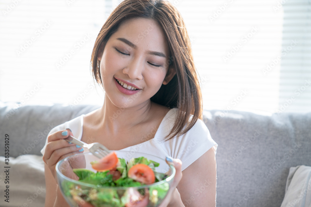 Young happy Asian woman eating healthy salad sitting on the sofa in living room.