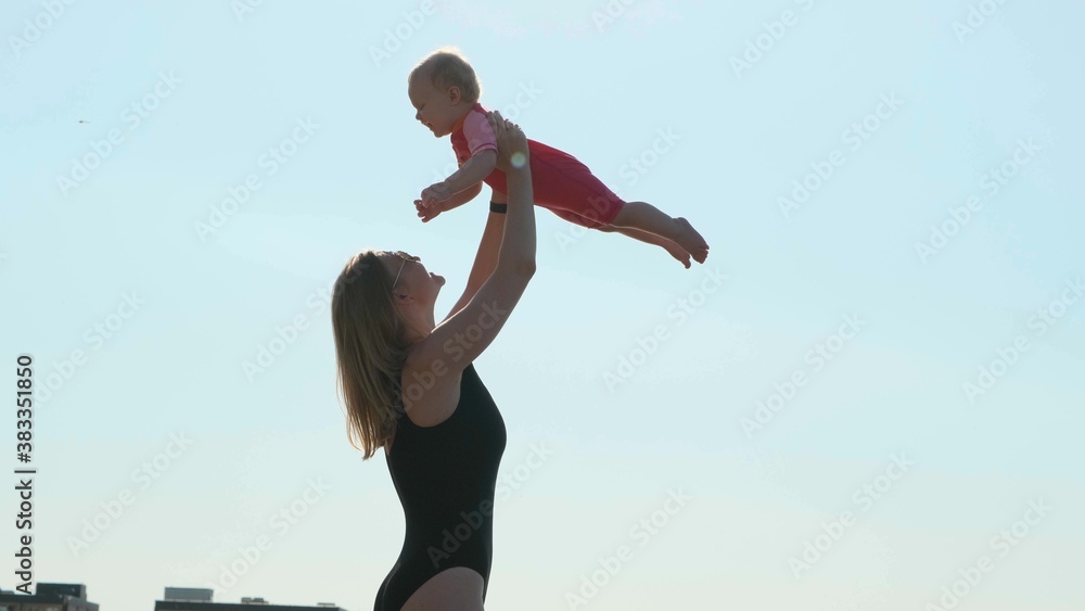 Obraz premium Sexy mother in black closed swimsuit on seashore or ocean throws her daughter up. Child in pink bathing suit. Nice view of happy family on vacation.