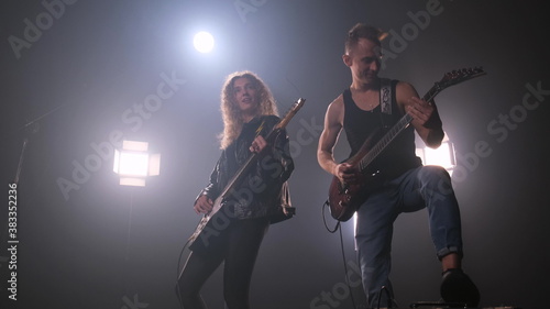 Two rock stars on stage. A lot of light on background. Guy in black T-shirt plays the electric guitar and woman with lush curly hair in leather jacket play too and enjoys music. fog around