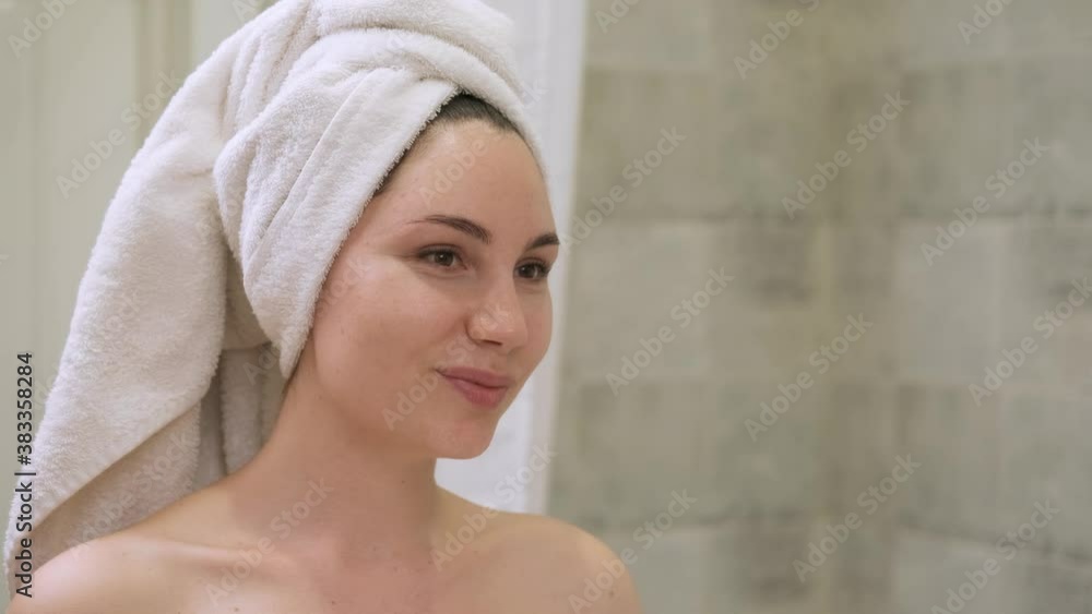 A young woman smiles and admires herself in the bathroom. Girl with a towel on her head after taking a shower. A woman looks in the mirror A young woman smiles and admires herself in the bathroom. Girl with a towel on her head after taking a shower. A woman looks in the mirror